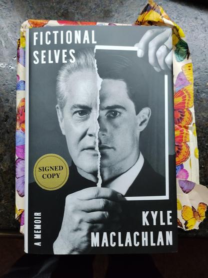 A photo of the book cover for Kyle Maclachlan's 'Fictional Selves', his memoir. It features Maclachlan holding a black and white photo of his face, as Agent Cooper circa 1990, torn in half vertically. We see half of his modern day face, and half of the Cooper face. There is a sticker on the cover with the words Signed Copy.