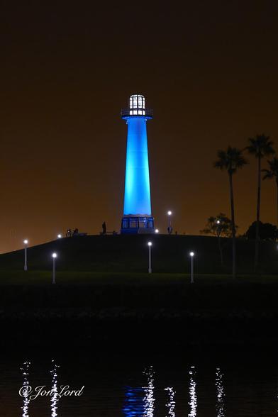 This is a night time colour photo in portrait format of a tall, illuminated lighthouse located on a small hill, harbour water in the foreground and a dark caramel coloured sky in the background. Long Beach, California (2012).
The dark waters of the harbour rise from the base of the photo, upwards to a little less than the one quarter up level, where the boundary between the water an distant shore is ill defined. The camera is located on the near shore, a few metres above the water and angled at the distant shore and lighthouse. The jet black water is rippled with the six long reflections from the small street lights on the shore. One further and shorter reflection is blue from the illuminated lighthouse. Above the shore and just less than the halfway up level is a path that slopes upwards gently from the right to the left. Along this path are four of the cylindrical poles with a light on top. Behind the path is the small hill with the lighthouse on top, in the centre of the photo and viewed from twenty-five metres away. The lighthouse is tall, cylindrical and tapers towards the white lantern at the top. The lighthouse is illuminated giving it a light blue appearance. Dominating the background and the upper half of the image is a dark caramel (nicotine?) coloured sky that graduates becoming ever darker the higher up in the photo.