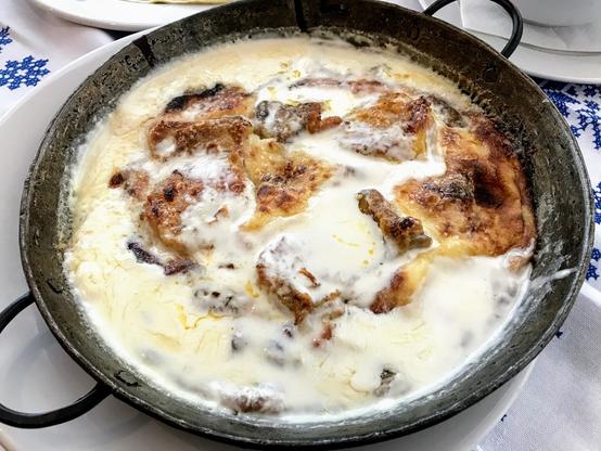 A rustic pan of browned bread pieces and chicken soaking in a creamy white sauce, served on a blue-and-white patterned tablecloth.