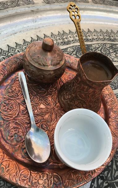 An ornate copper coffee set—a long-handled pot, lidded container, spoon, and white cup—rests on an embossed copper tray.