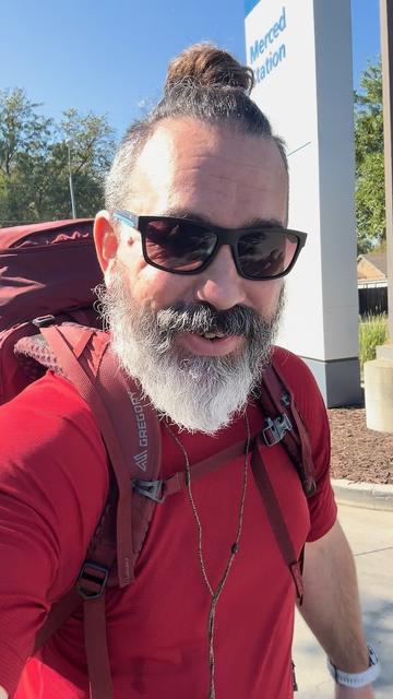 A bearded man wearing sunglasses, a red shirt, and a hiking backpack smiles outdoors beside a Merced Amtrak Station sign.
