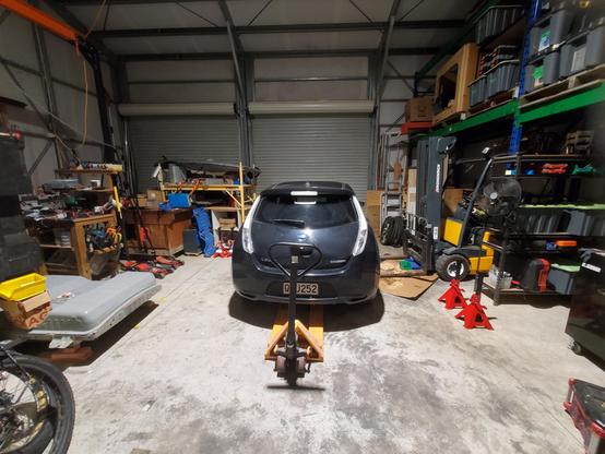 Photo inside a workshop. Junk around perimeter. Center is rear of black Nissan Leaf with a pallet truck under it. Left  is 40kWh battery. Right is forklift in a pile of absorbent sawdust. Also of note are 4 x 615mm tall axle stands. Did I mention that there's junk all around? Good. I'm working on reducing it...
