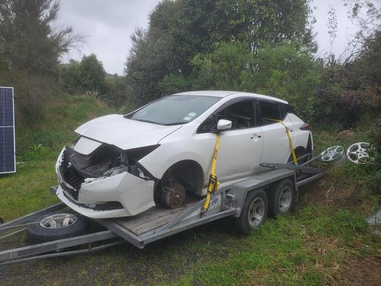 Sad looking white Nissan Leaf with no headlights, grille or wheels & bumpers hanging off. It's on a  trailer and is tied down through its open windows by yellow straps.