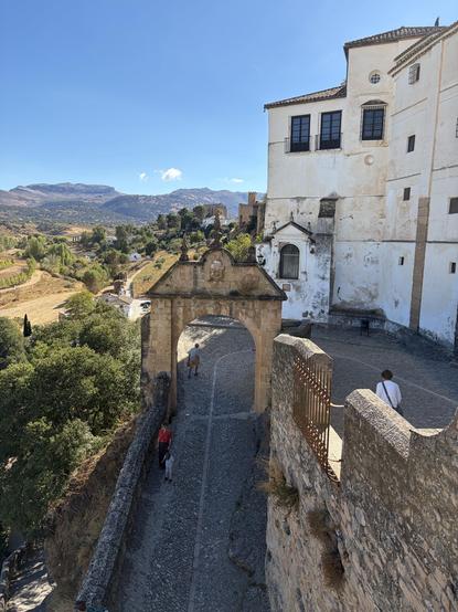 Looking down at a curved paved path that goes under a very old small city gate. There are old white buildings on one side and then the view drops away to lovely green and gold hills under a blue sky.