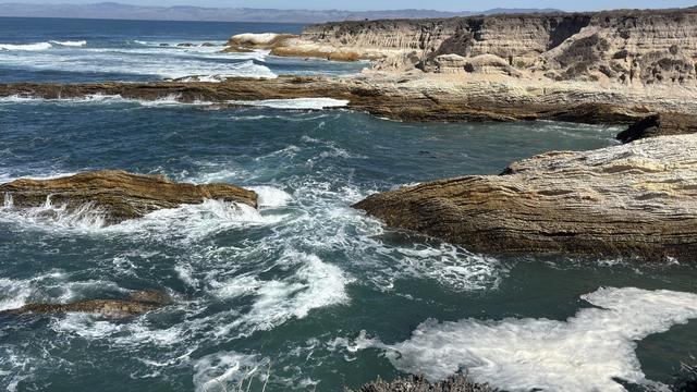 A scenic coastal view featuring rocky shorelines and crashing ocean waves. The water is deep blue, with white foam from the waves, and rugged cliffs are visible in the background.