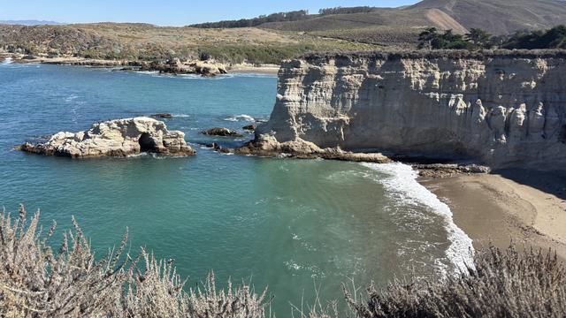 A scenic coastal view featuring rocky cliffs and clear turquoise waters. An arch-shaped rock formation is visible in the water, with a sandy beach nearby. The background includes rolling hills and greenery under a clear blue sky.