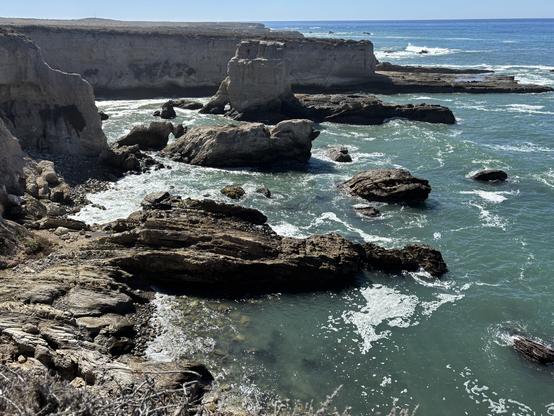 A coastal scene featuring rocky cliffs and waves crashing against them. The water is a mix of turquoise and deep blue, with foam on the surface. The coastline is rugged, with large boulders and a backdrop of steep cliffs under a clear sky.