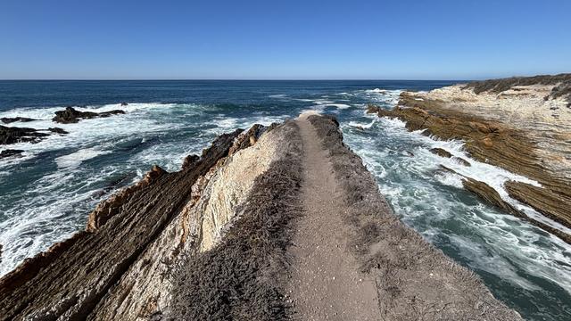 A coastal landscape featuring a narrow path bordered by rugged cliffs, with waves crashing against the rocks on both sides. The sky is clear and blue, complementing the vibrant ocean.