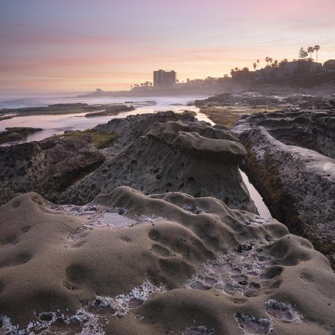A color photograph of rock formations along the coast of La Jolla, San Diego, California. Wispy clouds light up in beautiful colors with sunrise.