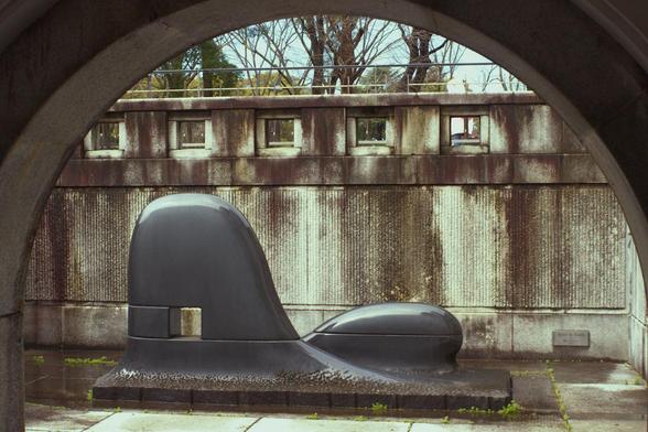 Abstract sculpture in some kind of old dark granite. Bulby shapes, two.  Old concrete wall slightly decorative with some appealing old waterstains. Framed by an arch.