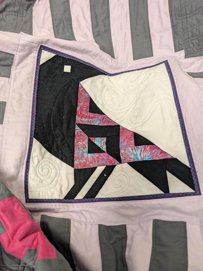 A quilt, featuring one of the blocks. It is a black crow that has a wing made of hot pink batik strips alternating with black. The colorful pieces are stitched in a colorful variegated thread. The crow has been outlined in black with some feather details added. The white background has pastel swirls coming in from the sides, like wind.