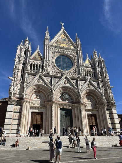 Facaden af Siena Duomo med uendelig mange detaljer i marmor, inklusive gargoyler af forskellig størrelse ude på hjørnerne. Et stort rundt vindue giver en mørkeblå kontrast til al det lyse marmor