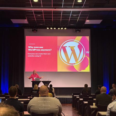 Sami presents on stage at a conference, with a large slide, poses a question, why, if people are using AI to create websites; audience members sit listening.