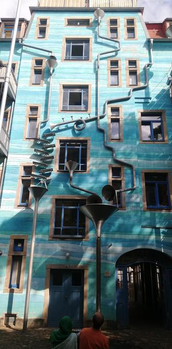 An einer in streifigem türkis bemalten Fassade mit kleinen schmalen und größeren normalen Fenstern laufen Regenrohre verwinkelt hinab. Sie fallen in Trichter oder gehen über Stufen nach unten und erzeugen dadurch Effekte.