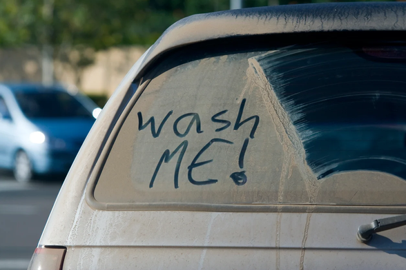 Closeup of the back window of a very dirty car.  Someone has written “Wash ME!” in the dirt with their finger.