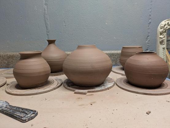 5 untrimmed tan stoneware clay wheel thrown vases drying on a table. They feel a little squat and heavy as there is excess clay at the base from when they were thrown.