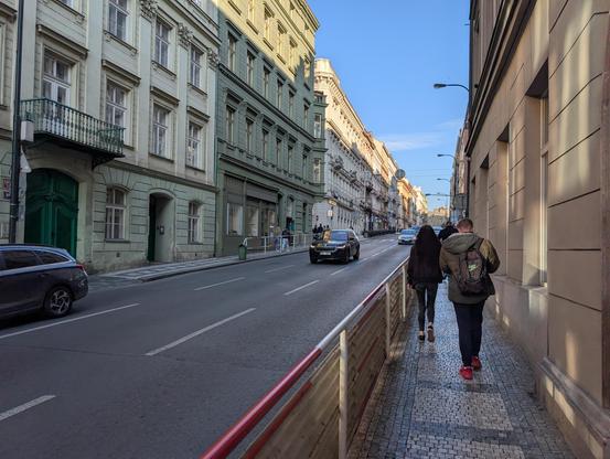 Narrow sidewalk with a solid fence separating it from a 3-lane unidirectional road, in a dense area with 5-floor buildings. No space for trees with all these cars of course.