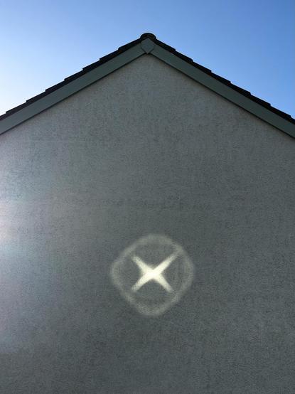 The gable end of a house with blue sky above. The wall has a prominent × with a rough circle around it created by reflected sunlight from a window opposite.