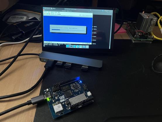 A small computer monitor displays a blue interface showing software installation progress. In front, an Arduino-like board is connected with a glowing LED. Cables are visible, along with a cooling unit and other equipment on a desktop surface.