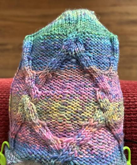 Eine begonnene gestrickte Socke.
Auf linken Maschen ziehen sich vier Stränge rechter Maschen in einem Diagonalen Muster entlang.
Bei jeder zweiten Kreuzung kreuzen sie sich zwei mal