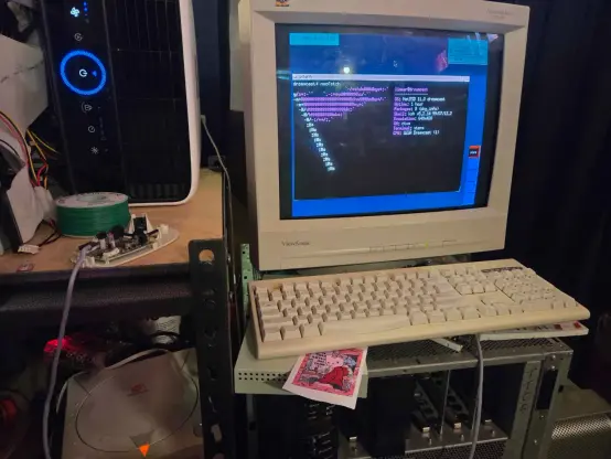 in my messy computer lab, a Sega Dreamcast sits on a shelf next to my SGI Onyx2, plugged into a ViewSonic CRT monitor. the dreamcast's keyboard and disassembled broken mouse are visible as well. the monitor shows the Dreamcast running NetBSD at its default ctwm desktop environment, showing a terminal with `neofetch` displaying system stats.