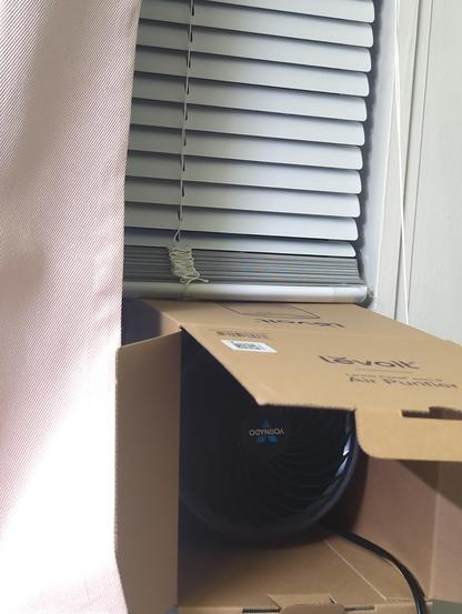 a box for a levoit air purifier that has been held in a sliding window by force of closing it on it. a smallish fan has been placed inside