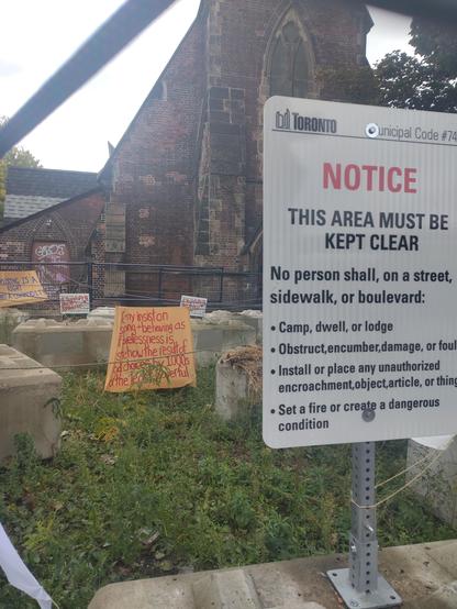Fenced-off church yard, with massive concrete blocks.

In the foreground, a city sign: "Notice, This area must be kept clear. No person shall, on a street, sidewalk or boulevard: Camp, dwell or lodge; Obstruct, encumber, damage or foul; install or place any unauthorized encroachment, object, article or thing; Set a fire or create a dangerous condition".

In the background, a painted sheet which says: "Too many insist on [?] and behaving as if homelessness is somehow the result of bad choices by 1000s of the least powerful."