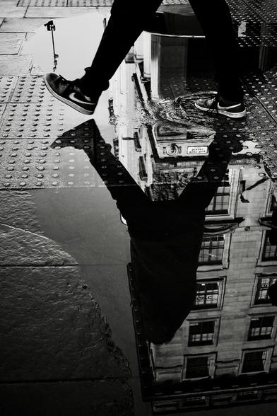 In a scene reminiscent of Henri Cartier Bresson's classic and rightly-famous 'Decisive moment' photograph, a man steps over a puddle of rainwater at the edge of a street where the ground is studded with metal bolts to allow blind people to know that they are approaching the edge of the pavement. The puddle is so large that it is clearly reflecting a tall building nearby. We can only see the subject's lower legs but his silhouette is shown in full in the reflection.