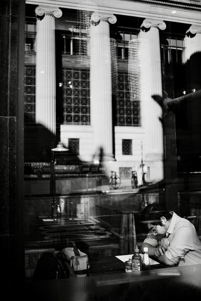 A man sits in a window seat at a restaurant with his coat and bag on the opposite side of the table. His arms are folded and rested on the table as he reads something, possibly a menu, while in the tall window the reflection of a brightly-lit classical building across the street dominates the upper two-thirds of the image.