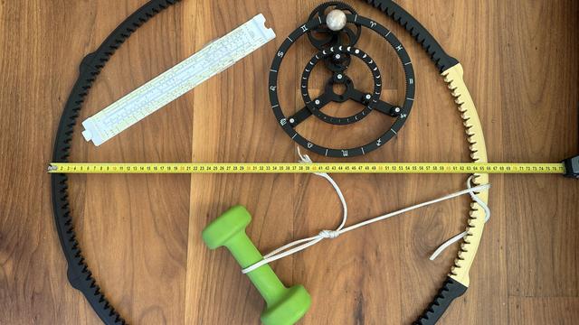 a large 3d printed ring gear with a tape measure across it, a 2kg hand weight tied to it, a sliderule and a smaller version of the orrery