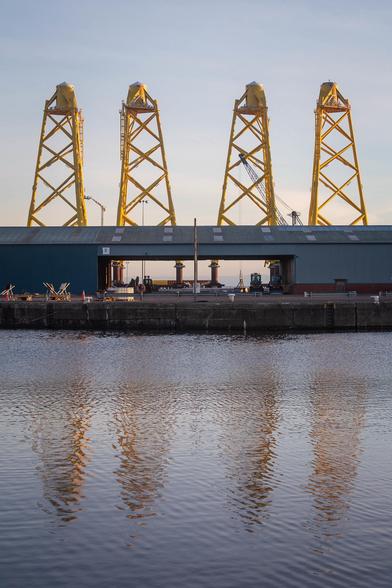 A huge shed sits on the dockside. Behind it, towering over everything else, are four enormous yellow structures. They’re jacket foundations, soon to be shipped off and submerged under the North Sea to support wind turbines.