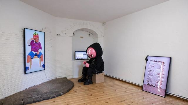 A black-clad character sculpture with a large pink, cartoon-like head sits hunched forward on a small wooden cube in a white-walled gallery. The head features a white social media symbol and large cartoon eyes. The figure wears a black hooded outfit, layered silver chains, black trousers, and chunky black boots, looking down at its hands. A vertical screen on the left displays a portrait of a person wearing a colorful striped sweater, denim shorts, white socks, and blue shoes. A second framed screen leans against the wall on the right, showing a pale pink and white abstract image. A small computer monitor is visible in an arched alcove behind the seated figure.