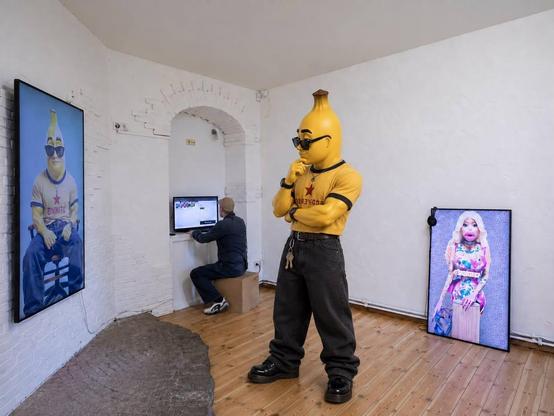 A banana-headed character sculpture stands in the foreground of a white-walled gallery, wearing black sunglasses, a yellow cropped T-shirt with a red star and lettering, loose black trousers, and chunky black boots. The figure poses with one hand on its chin and the other arm folded across its body. A vertical screen on the left displays a seated portrait of the same character in a yellow shirt and blue trousers. Another screen on the right shows a colorful portrait of a blonde female character with exaggerated features. In the background, a visitor sits on a wooden cube and operates a computer in a shallow arched alcove. The room has exposed white brickwork, wooden flooring, and a rough, dark stone-like feature along the base of the left wall.