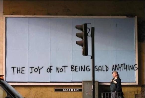 Photo of a person walking along a street past an empty billboard covered in white paper. Graffiti on the white paper reads, "the joy of not being sold anything."