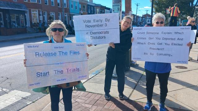 People holding 3 signs:

Release the files, start the trials

Vote early by November 3, Democrats over dictators

Could someone explain which crimes get you deported and which ones get you elected president? It's so confusing