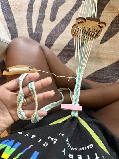 A photo from my POV, sitting cross-legged on the floor, weaving. My hand is holding a woven strap while using a small wooden loom with warp threads attached to my waist. The threads are white and turquoise and the band is white with a turquoise chain link pattern going down the center. The background features a zebra-striped rug.