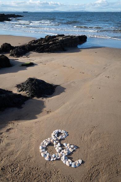 An ampersand formed of bits of broken white shells, on a plain sandy beach. Some rocks and the blue water of the Firth Of Forth are visible in the background.