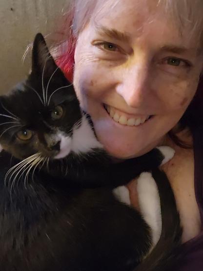 A black and white tuxedo kitten curled up around the neck of a white lady with grey and red hair. Both are looking up into the camera.
The woman is smiling, and the kitten is purring.