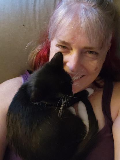 A black and white tuxedo kitten curled up around the neck of a white lady with grey and red hair, who's wearing a purple tank top. The woman is smiling and looking up into the camera.
The kitten is resting her head on the woman's chin.