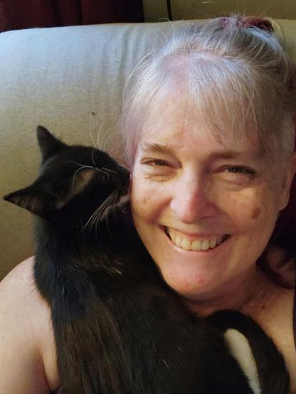 A black and white tuxedo kitten curled up along the neck and shoulder of a white lady with grey and red hair.
The woman is smiling and laughing as the kitten is licking her cheek.