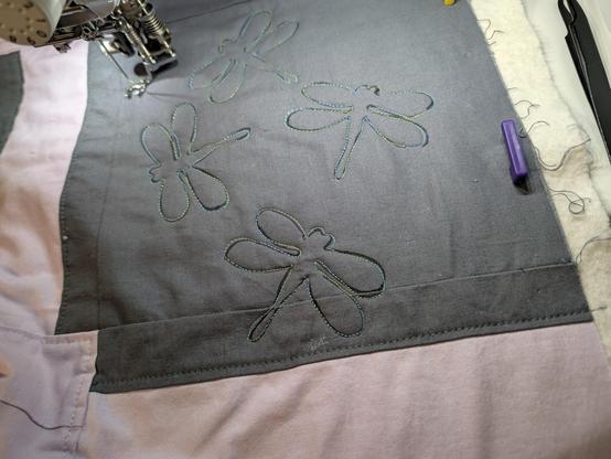 Four smaller dragonflies in shiny green-to-blue variegated thread on a grey background block on the edge of a quilt.