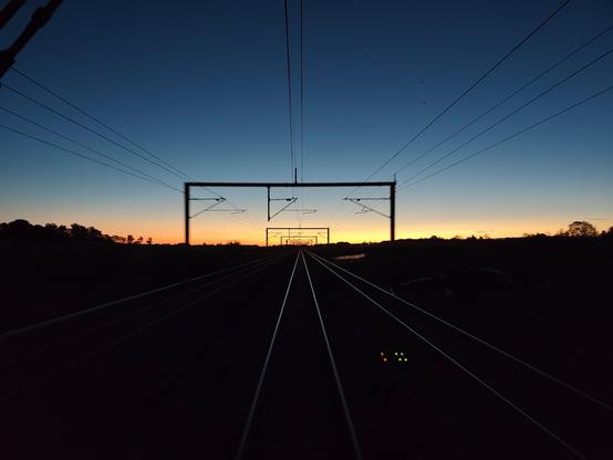 View out the rear of an IC3 train, travelling west.
Sun is barely rising, with a clear sky. Overhead wires and masts cast dark contours.