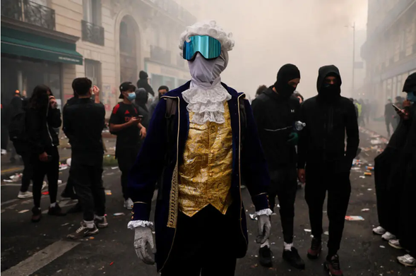 Foreground is a protester in 18th century costume: a periwig, white shirt with frilled collar, golden yellow silk waistcoat and brocaded acket.  Their face is completely covered in white material, with  safety goggles over thet op.  Behind them is a row of protesters in black, outlined against a pall of smoke from tear gas cannisters.

Picture: AFP