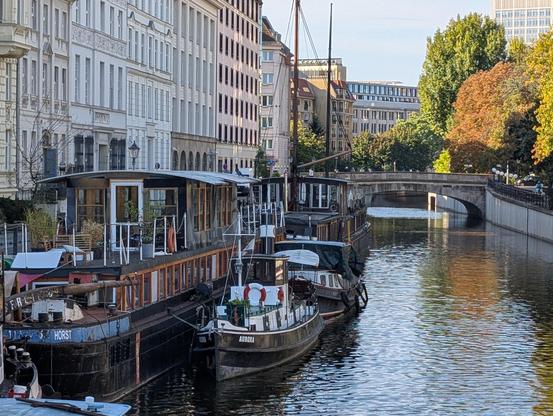 Two old boats on a canal. In the background, tall buildings and a small bridge.  A few fall colored trees. Light on the water.

Photo: Brad Rubenstein (2026)