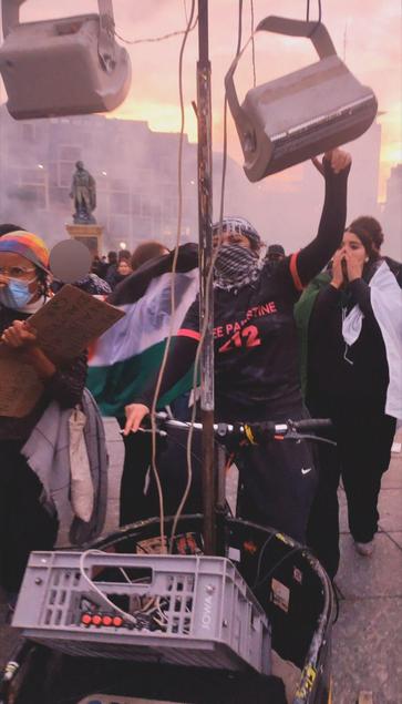 une jeune femme qui pilote le queergo avec la rête cagoulée dans un keffieh noir et blanc et un maillot de foot free palestine, d'autres filles sont là autour en mouvement avec un drapeau algérien et un drapeau palestinien