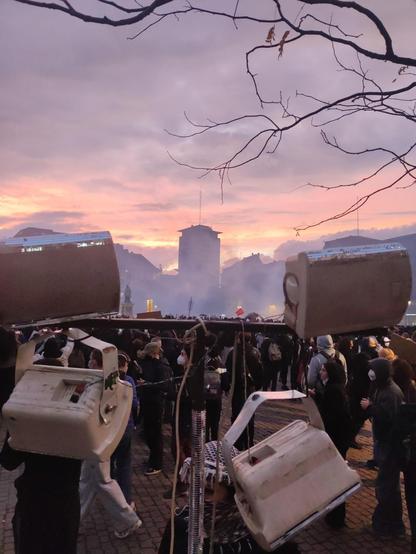 la place kléber avec une foule au coucher du soleil, embrumée par les gaz lacrymo avec les enceintes du queergo en avant-plan dont une pendouille à un rizlan car sa monture a cédé, des branches d'arbre en surplomb, la silhouette d'une tour au fond trône au centre de l'image.