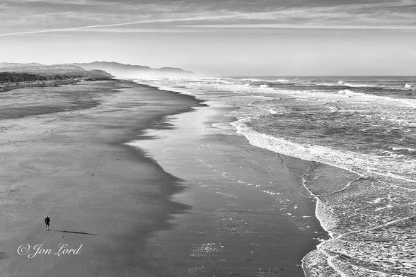This is a black and white photo in landscape format of an elevated view along a nearly deserted and wide beach that stretches for miles with the sea on the right side and a single man walking in the lower left corner. Ocean Beach, California (2017).
The camera is located thirty metres above the beach and is aligned with the beach on the left side of centre and the sea on the right. The beach and ocean rise upwards from the base of the photo to a little over the three-quarters up level. The beach is wide, at least one hundred metres from the shore to the water, perhaps more. The beach has ragged streaks extending along parallel to the water, indicating the high water mark and the point where the surf from the breaking waves is reaching: the latter has a shiny surface from the recent passage of water. From the centre to the right edge of the image is the Pacific Ocean. The beach must continue out into the sea with a gentle and shallow gradient since there is one hundred metres of surf before the breaking waves are reached along the right edge of the photo. In the lower left corner is a single man, not much more than a small speck, walking along the beach towards the camera. In the distance, in the upper left corner is a ridge of hills that stretches from the left towards the centre of the image. Above is a near, clear and cloudless sky with just the smallest wisp of cirrus high clouds and two contrails.