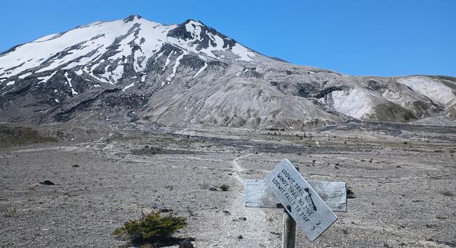 A trailsign is askew on the post but says Loowit trail. The trail is a dusty track across a barren plateau ahead, from which rises the bulk of Loowit herself, also known as Mount Saint Helens.
