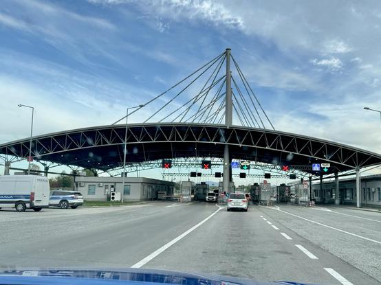 A large concrete roof, strapped by steel cables to a set of concrete pillars, stretches over multiple lanes of road, with little booths in between. Two lanes have signs over them with green arrows, three have red crosses.
