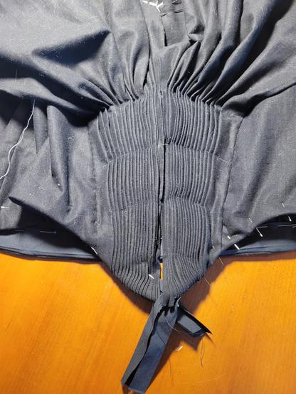A detail about of a black dress bodice on a wood table. 

The bodice is black linen, and closes center front,  meeting in a V shape at the waist. The center front panels is intricately pleated into tiny knife pleats (16 on each side) releasing at the bust to loose, soft folds.